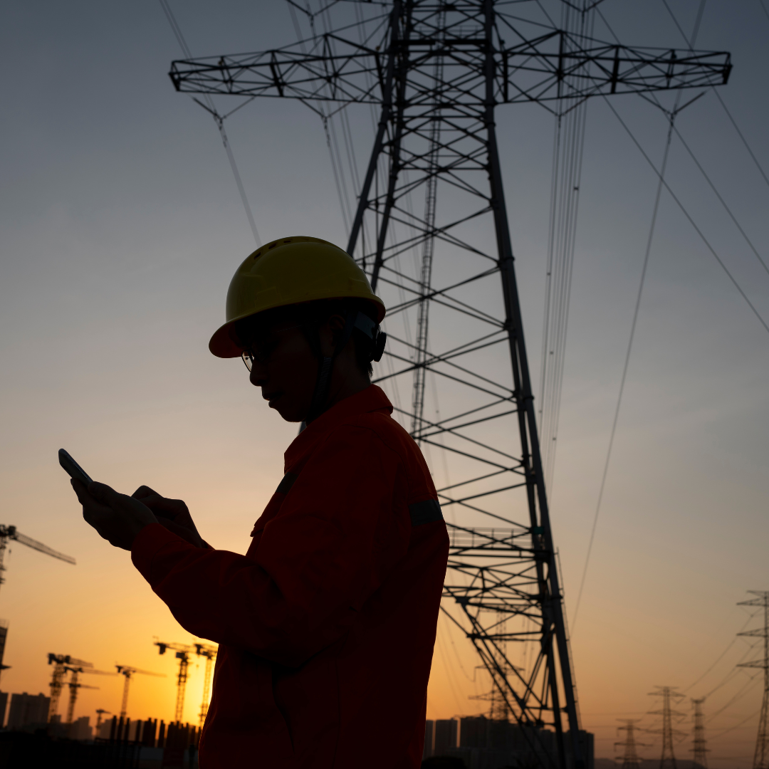 A power system consultant silhouetted by the sun, standing near an electricity pylon.