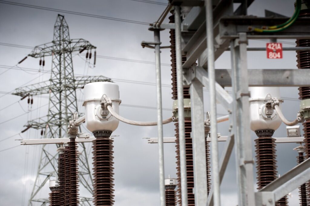 High-voltage substation insulators and instrument transformers with a transmission tower behind, under an overcast sky.