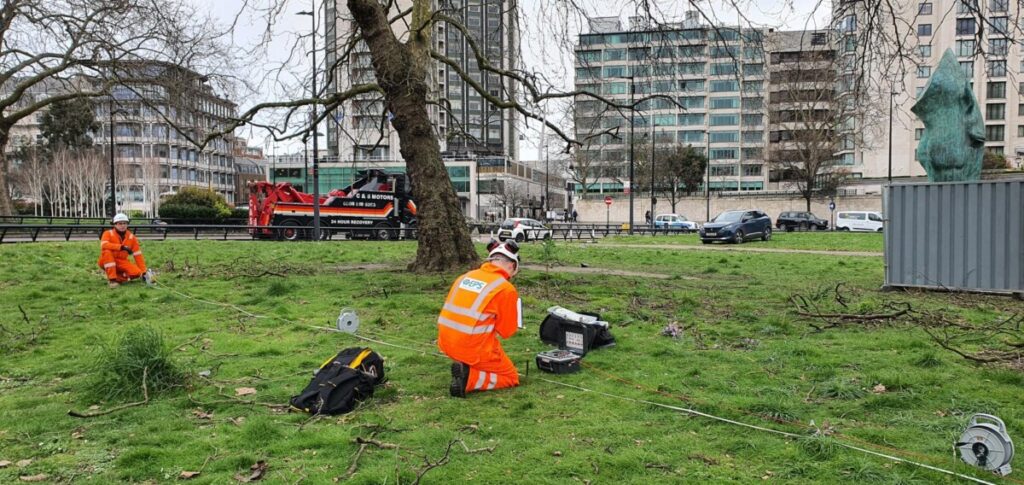 Two EPS engineers in branded orange hi-vis kneeling on grass conducting earthing site inspections.